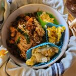 An image of teriyaki fried noodles presented in a ceramic bowl nested in a basket. There is a towel wrapped around the bowl. Inside the bowl, scrambled egg curds are separated from the fried noodles in silicone muffin cups.