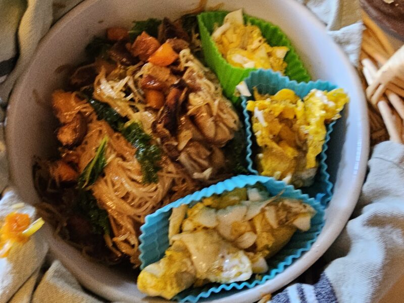 An image of teriyaki fried noodles presented in a ceramic bowl nested in a basket. There is a towel wrapped around the bowl. Inside the bowl, scrambled egg curds are separated from the fried noodles in silicone muffin cups.