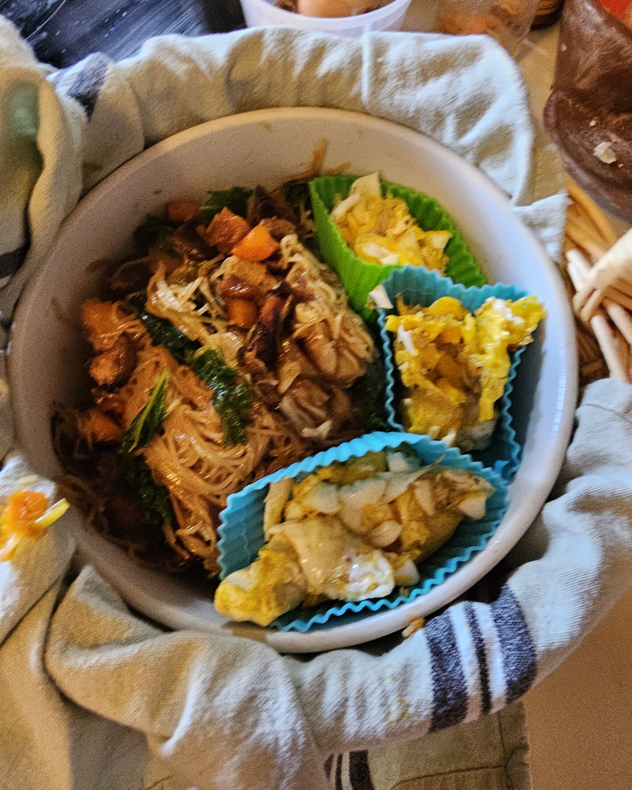 An image of teriyaki fried noodles presented in a ceramic bowl nested in a basket. There is a towel wrapped around the bowl. Inside the bowl, scrambled egg curds are separated from the fried noodles in silicone muffin cups.