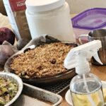 A crop of an image of the kitchen table, the apple crisp pie is surrounded by various baking ingredients, vegetables, and pot pies in the process of being prepared.