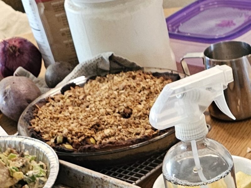 A crop of an image of the kitchen table, the apple crisp pie is surrounded by various baking ingredients, vegetables, and pot pies in the process of being prepared.