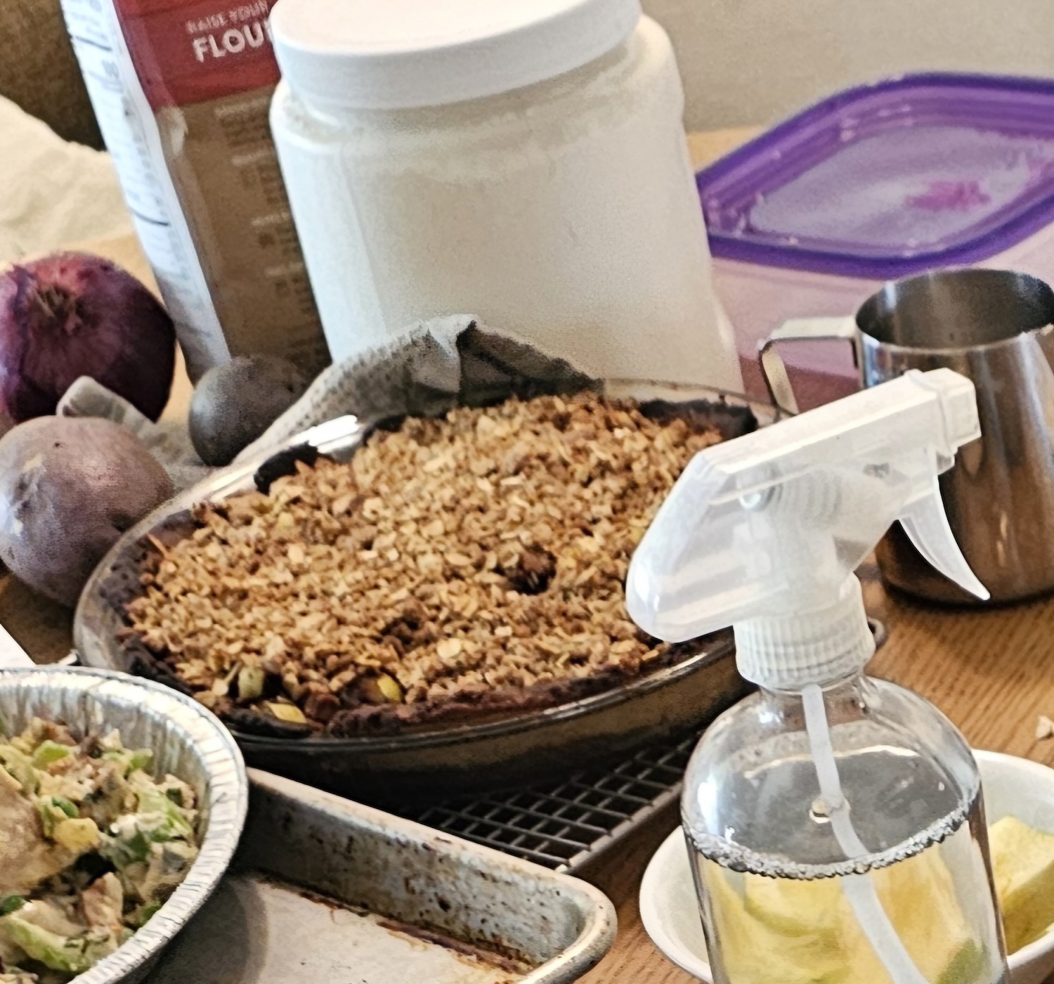 A crop of an image of the kitchen table, the apple crisp pie is surrounded by various baking ingredients, vegetables, and pot pies in the process of being prepared.